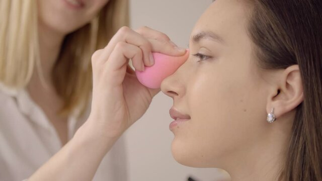 Blonde make-up artist applies face powder on a model's face. Close-up of a MUA using pink sponge for putting on a cosmetic product. High quality 4k footage