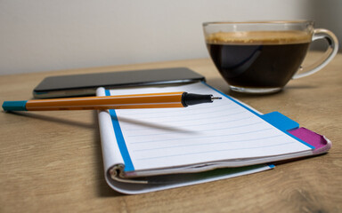 A notepad with white pages and horizontal lines with an orange pen with no lid on top with a cup of coffee and a cell phone in the background on an office desk referring to the home office.