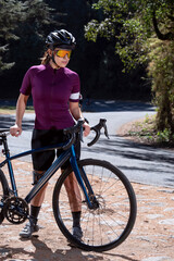 Chica con su bicicleta de pista, usando casco y lentes de sol, junto a la carretera en medio del bosque