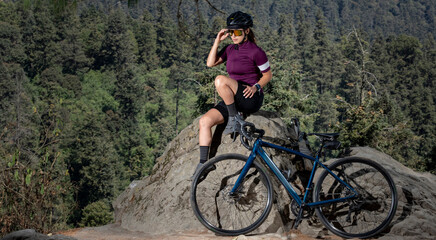 Plakat mujer latina con bicicleta descansando en el bosque, con un paisaje de arboles al fondo. Concepto deporte al aire libre