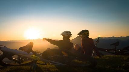 Girls sitting on mountain with bycicles looking at sunset and talking. Enjoying in beautiful nature.