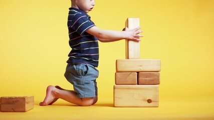 Cute 4 years old boy is playing with big wooden bricks, building a tower. Studio, yellow background