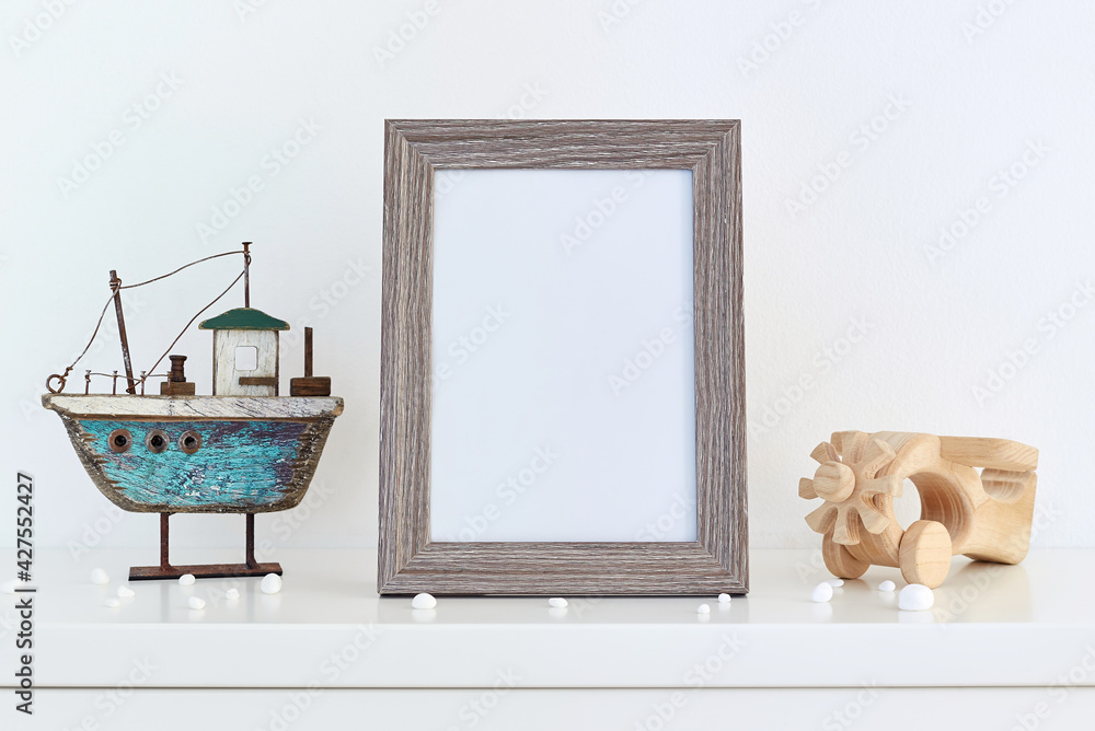 Wall mural Mock up picture frame on white shelf with wooden boat and wooden airplane against white wall; portrait orientation; stylish interior mockup background with modern accessories