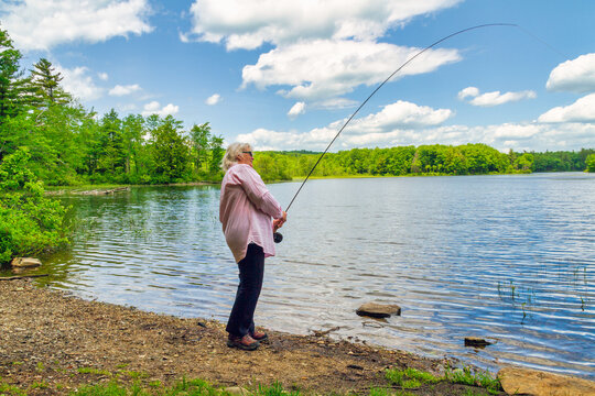 Female senior citizen spending a beautiful summer day fly fishing in the lake. Surrounded by green trees, blue sky and puffy clouds.