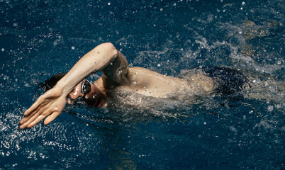 Obraz na płótnie Canvas Caucasian athlete-swimmer crawls in the blue water. Portrait of a young male triathlete swimming in swimming goggles. Triathlon training concepts for triathletes