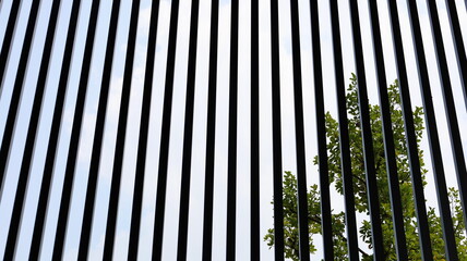 Slat shading light. Black silhouettes of metal lattice fences adorn vertical modern houses or buildings with green plant background and blue sky. Selective focus