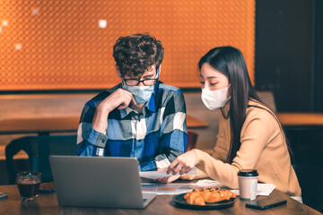 young asian woman and caucasian man Business people Working discuss finding new strategy solution.People putting  protective face mask discussion business plan using laptop at cafe.