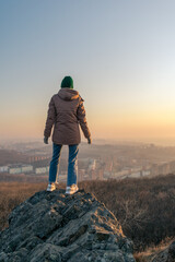 A girl in a jacket stands on a rock and looks at the city from a height during sunset