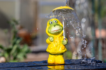 frog figure with rain jacket stands in the rain in spring and is protected by a yellow rain...