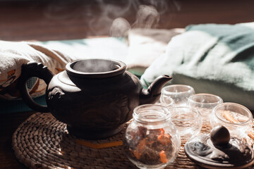 Hot tea in a clay teapot, rising steam, mugs on a tray inside the room. Morning tea ceremony concept. Close-up, selective focus