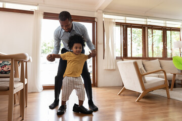 Smiling African little child having fun stepping with his dad shoe in living room at home. Happy...
