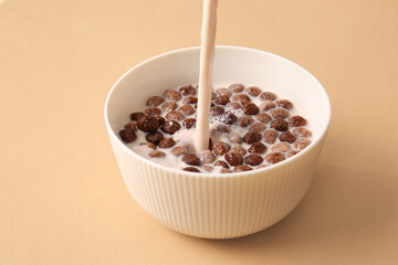 Pouring of milk into bowl with chocolate corn balls on color background