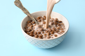 Pouring of milk into bowl with chocolate corn balls on color background, closeup