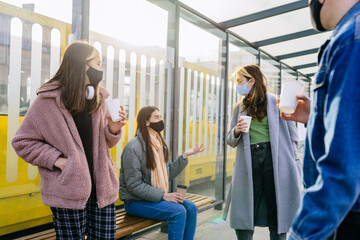Four people waiting on bus stop for a public transport. Group of friends millennial man and women waiting drink coffee to go and talking at bus stop commuting