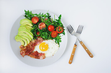 tasty breakfast - fried egg and bacon served with arugula tomato salad and fresh avocado on white plate