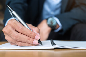 Businessman in suit writing something on notebook in office or cafe, hand of man holding pen with signature on paper report. business concepts