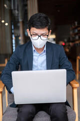 young Businessman in suit wearing surgical face mask and using laptop, man typing keyboard computer notebook in office or cafe. Covid-19 Pandemic, technology and New normal concept