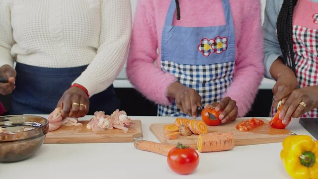 Close-of black females hands slicing chicken and fresh organic vegetables with knives on wooden cutting board, preparing meal together while united family enjoying cooking in domestic kitchen