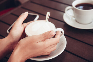 Cup of cappuccino with latte art in beautiful female hands and a cup of black aromatic coffee on wooden table. White ceramic cup of coffee. Open-air rustic morning coffee