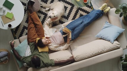High angle shot of woman lying on sofa, reading book and petting dog as her Afro-American husband browsing the web on smartphone while spending time together at home - Powered by Adobe