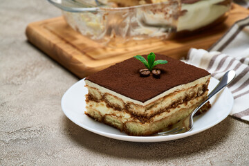 Traditional Italian Tiramisu dessert in glass baking dish and portion on grey concrete background