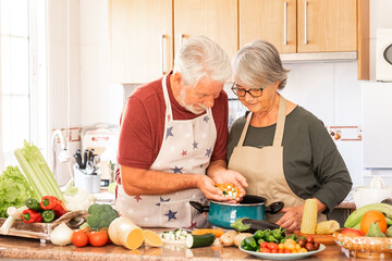 Two smiling seniors in chefs aprons prepare vegetables for a soup together in the home kitchen. Joyful elderly couple who appreciate a vegetarian lifestyle and enjoy their retirement