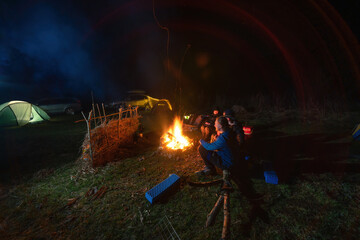 Group of tourists relaxing near campfire after a car trip, Lighting tents, camping equipmrnt. Night spring time. Back view.