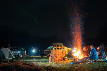 Group of travelers sitting near bonfire, cars and camp tent under beautiful night sky. View of blue starry sky over meadow with travelers near campfire.
