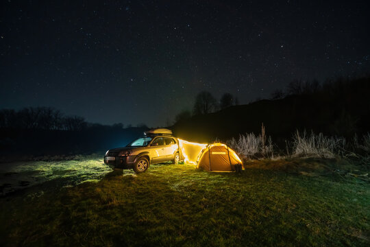 Night Landscape With Illuminated Tent And 4wd Car, Lights And Stars In The Background. Spring Camping. Tourism And Vacation. Roof Rack, Car Luggage Box