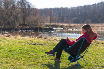 outdoor portrait of woman sitting in folding chair with smartphone near the river and forest. day time. Travel equipment. Copy space