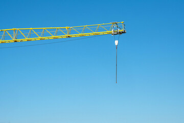 A yellow construction crane made of steel pipes with a hook on which hangs a chain against a blue sky.