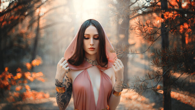 Portrait of beautiful woman fantasy elf in autumn forest. Dark trees background, fog park. Girl princess in orange peach color silk dress, hood on head. Sexy fashion model dreaming face brunette hair.