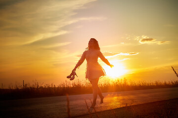 Beautiful sexy girl in pink dress and with high-heeled shoes in hand walking on the asphalt highway during sunset and field with grass on the side of the road
