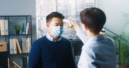 Rear of female worker standing in cabinet at work using wireless infrared thermometer measuring temperature of mixed-races male colleagues. Businessmen in medical masks working in quarantine, close up