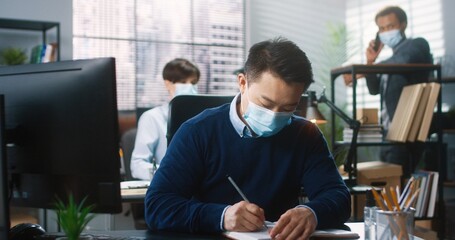 Portrait of Asian male worker sitting at desk in cabinet typing on computer and writing in planner, work in coronavirus quarantine. Colleagues on background. Man in medical mask work in company office