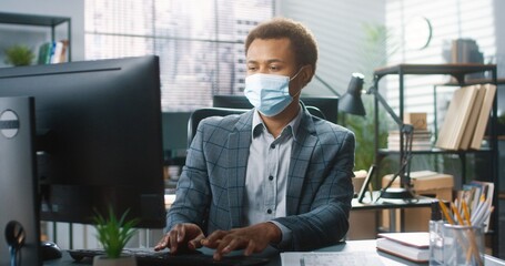 Portrait of young African American male worker in medical mask sit at workplace at desk and typing on computer while coughing and having headache. Coronavirus pandemic, covid-19 symptoms. Work concept
