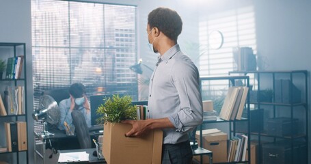 Portrait of young African American male worker in medical mask standing in cabinet holding carton box in hands with his stuff and looking at camera, fired from work, new employee at office