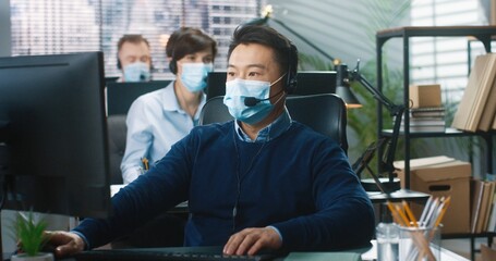 Portrait of Asian happy male call center worker in medical mask sitting at desk at company cabinet...