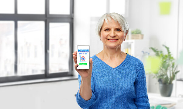 technology and health care concept - happy smiling senior woman holding and showing smartphone with international certificate of vaccination on screen over office background