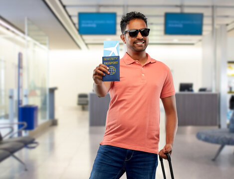 Safe Travel, Tourism And Health Care Concept - Happy Smiling Young Indian Man With Air Ticket And Immunity Passport Over Airport Background