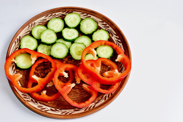On a clay plate are sweet bell peppers and a fresh green cucumber.