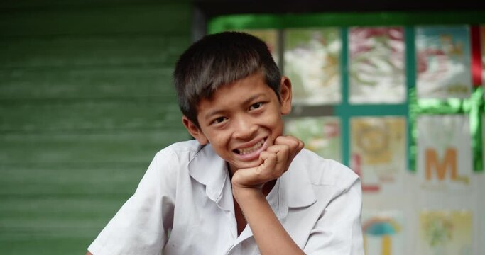 Slow motion of smiling poor Asian elementary school student boy in white uniform rest his chin on his hand and
standing in font of old green wooden wall of his school at the countryside.