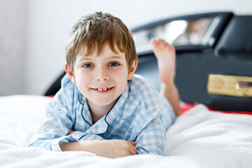 Adorable happy little kid boy after sleeping in his white bed in colorful nightwear. School child celebrating pajama party and looking at the camera. Funny happy child playing and smiling