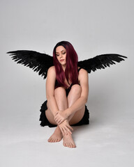 Full length portrait of a red haired  girl wearing black dress and feather angel wings.  Sitting...