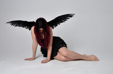 Full length portrait of a red haired  girl wearing black dress and feather angel wings.  Sitting pose against a studio background.
