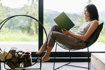 Beautiful stylish woman relaxing with book in modern chalet with view on mountains. Leisure time