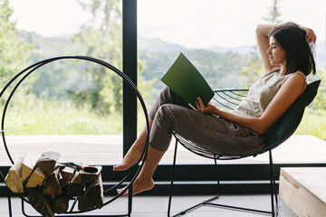 Beautiful stylish woman reading book on chair at firewood at window with view on mountain hills