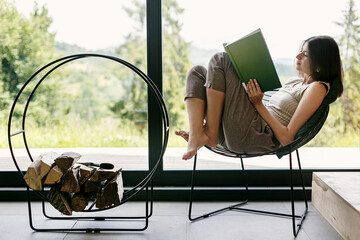 Beautiful stylish woman reading book on chair at firewood at window with view on mountain hills
