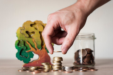 Una persona coloca con la mano una moneda encima de una pila de monedas, un bote de cristal lleno de monedas y un árbol de madera