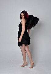
Full length portrait of a red haired  girl wearing black dress and feather angel wings.  Standing...